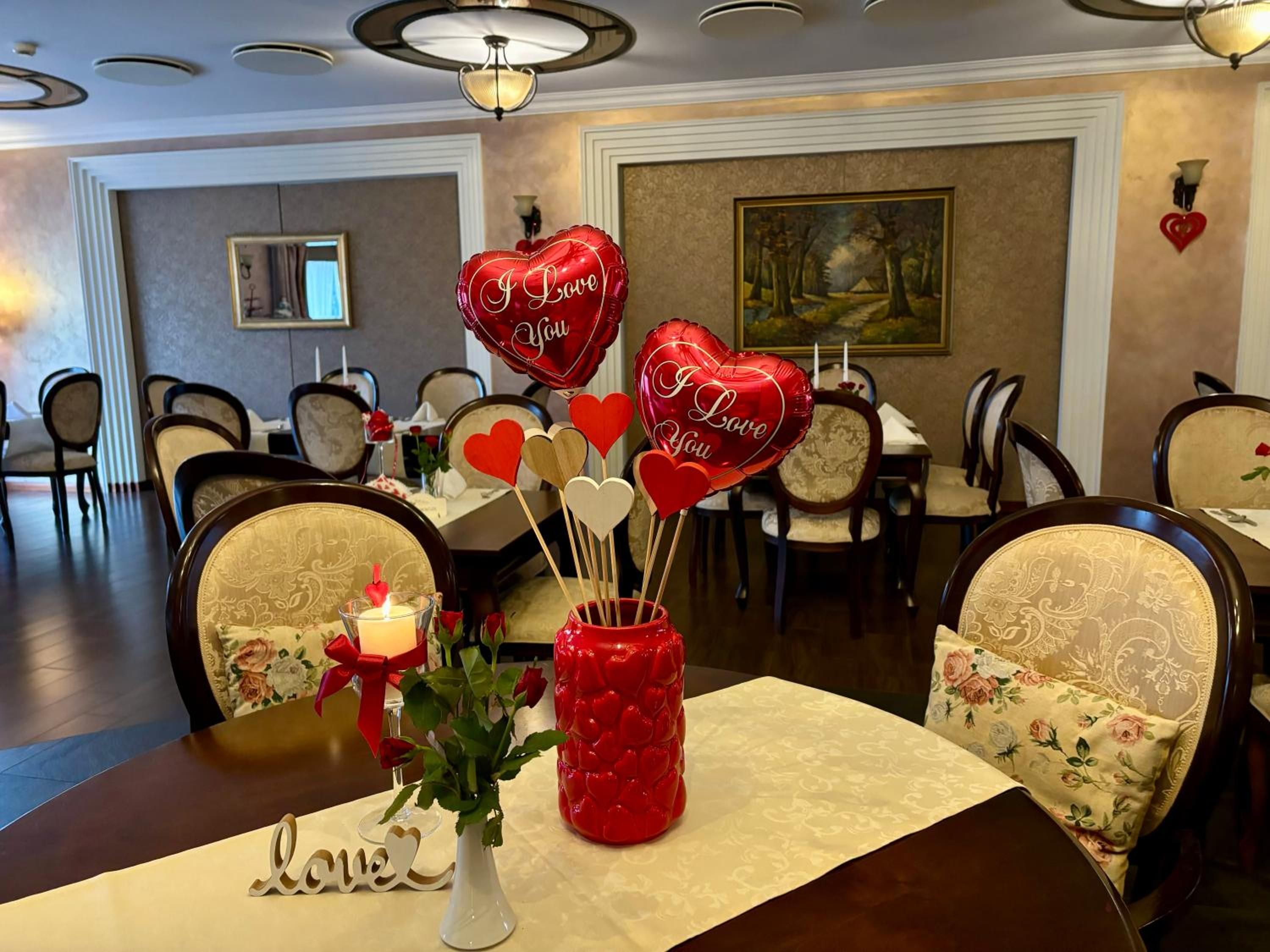 A dining table set with a candle, a vase with roses, heart-shaped balloons, and a decorative sign with the word 'love'.