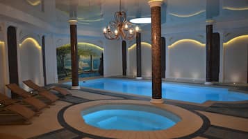 An indoor swimming pool with a jacuzzi, lounge chairs, and decorative columns.