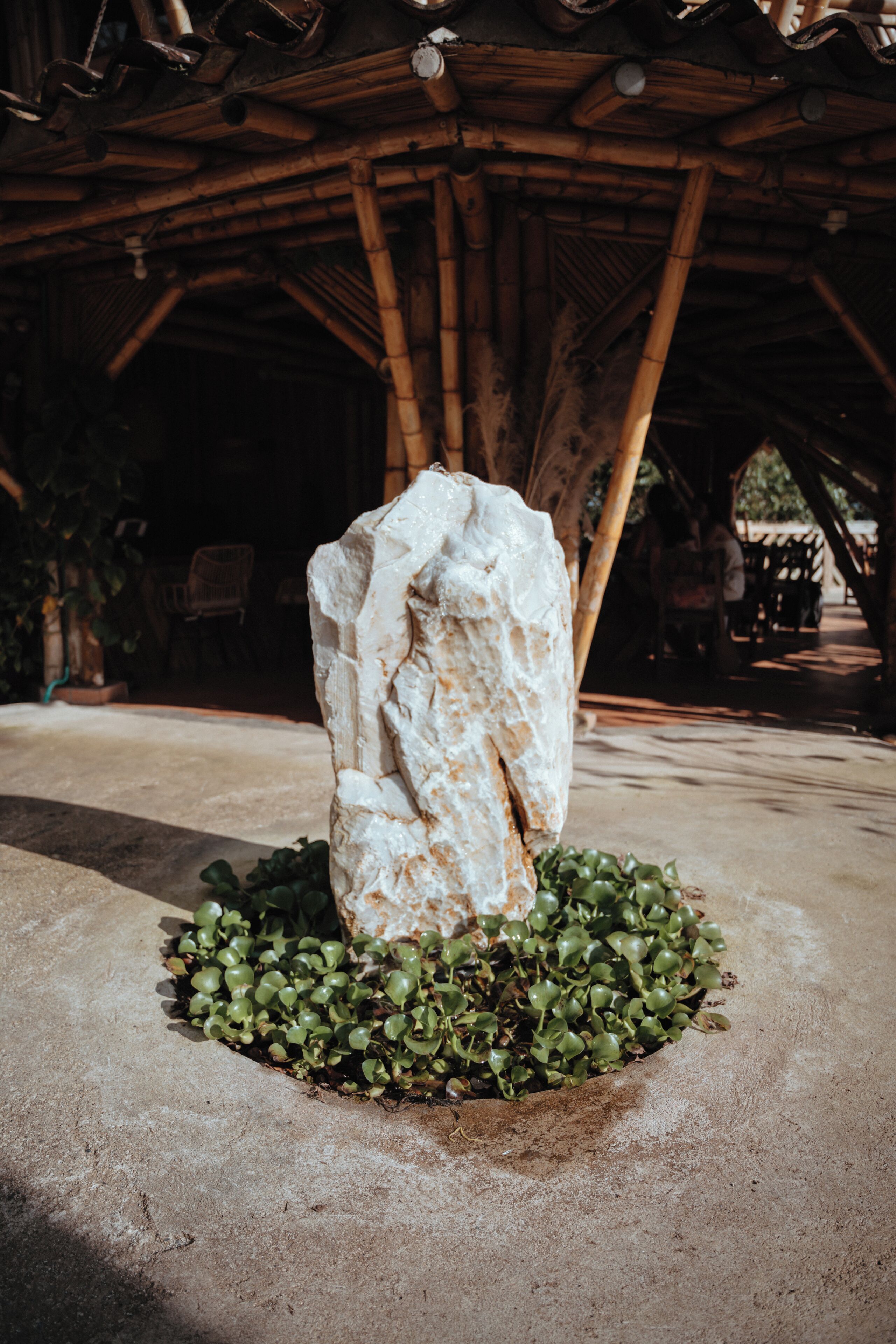 Una escultura de piedra en una jardinera circular con follaje verde, ubicada en un espacio exterior con una estructura de bambú al fondo.
