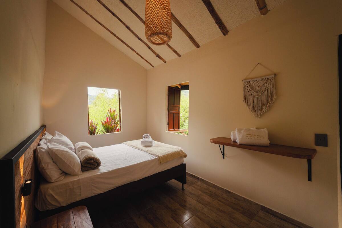 A bedroom with a bed, two windows, a wooden bench, a shelf with towels, and a macrame wall hanging.