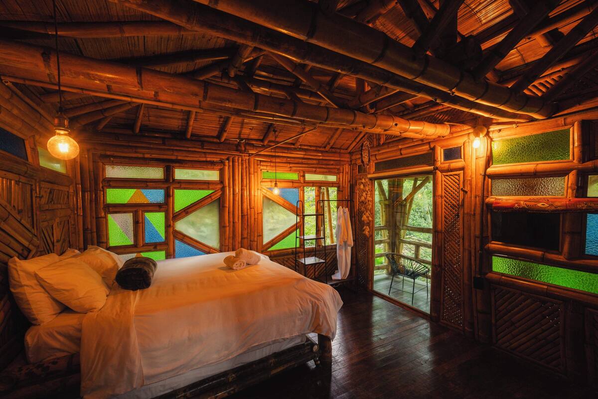 A wooden interior with a bed, a window with green glass panes, and a view of the outdoors.