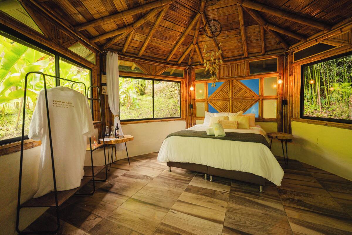 A room with a bed, a table with a towel, and a window with a view of greenery.