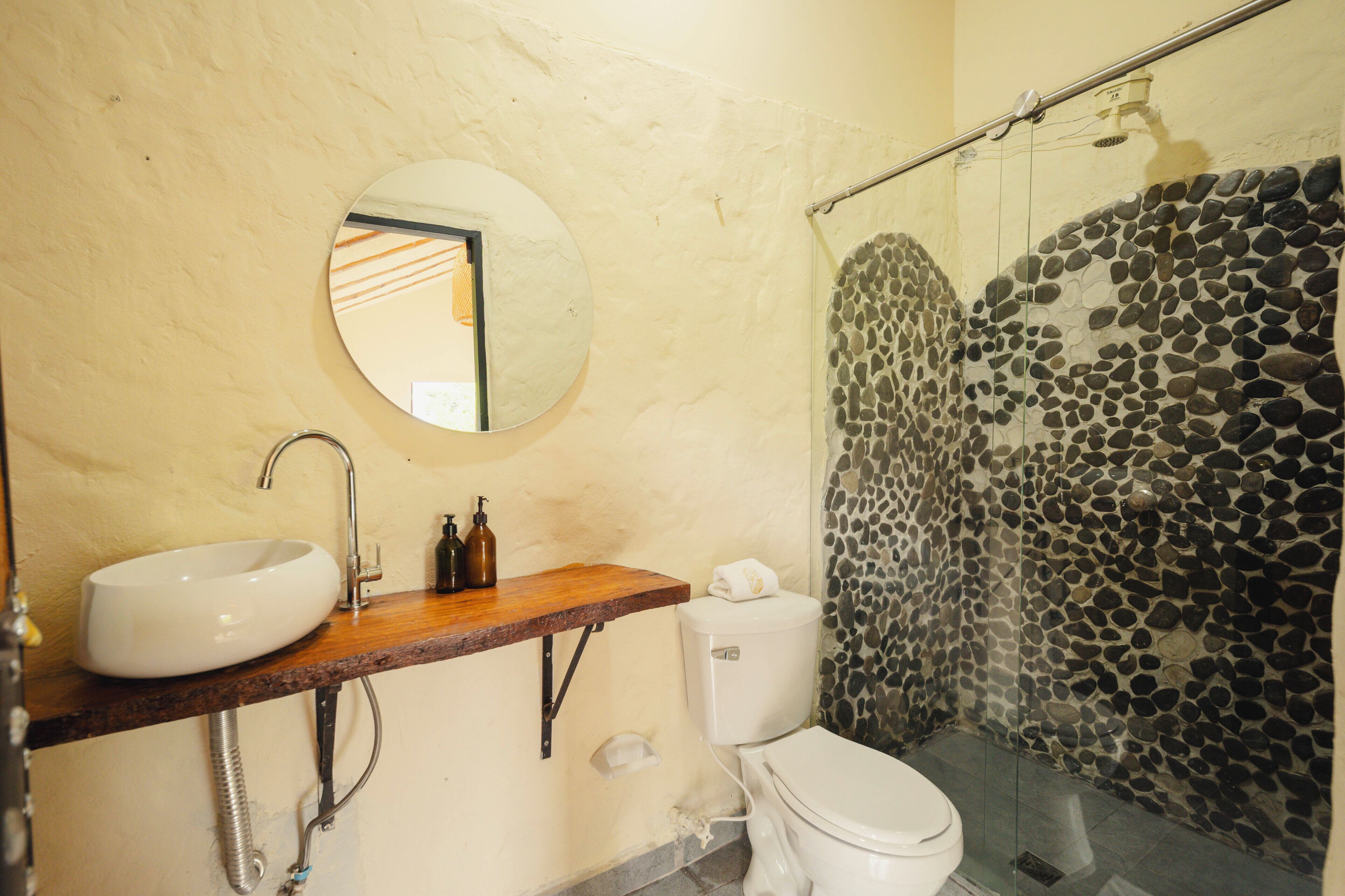A bathroom with a round mirror, a white sink, a wooden countertop, a toilet, and a glass-enclosed shower with a stone wall.