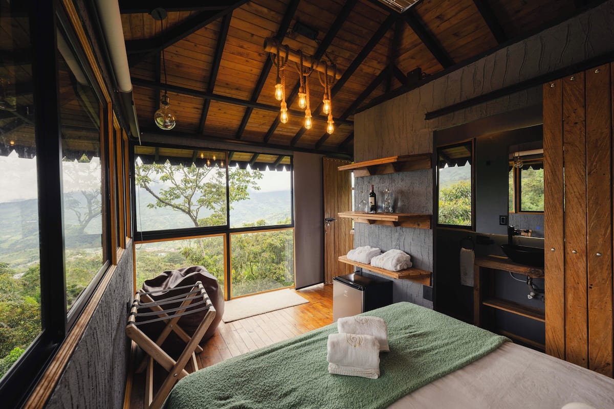 A bedroom with a large window, wooden beams, a bed with a green towel, and a small desk.