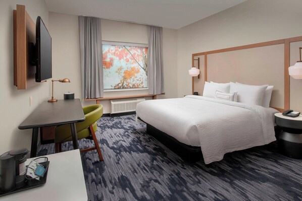 Habitación de hotel con cama, escritorio con silla, televisor montado en la pared, ventana con cortinas y vista a hojas otoñales.