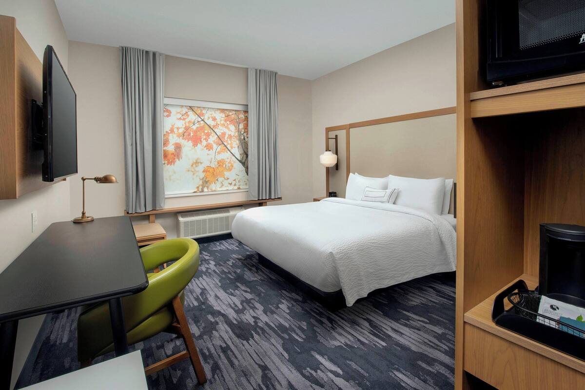 Ein Hotelzimmer mit Bett, Schreibtisch, Sessel, Fernseher, Fenster mit Vorhängen und einem Gemälde.