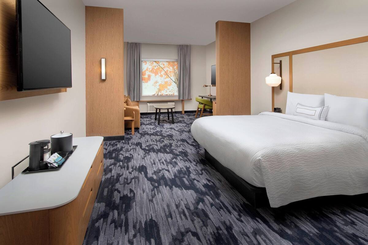 Ein Hotelzimmer mit einem großen Bett, einem Schreibtisch mit Stuhl, einem an der Wand befestigten Fernseher und einem Fenster mit Vorhängen.