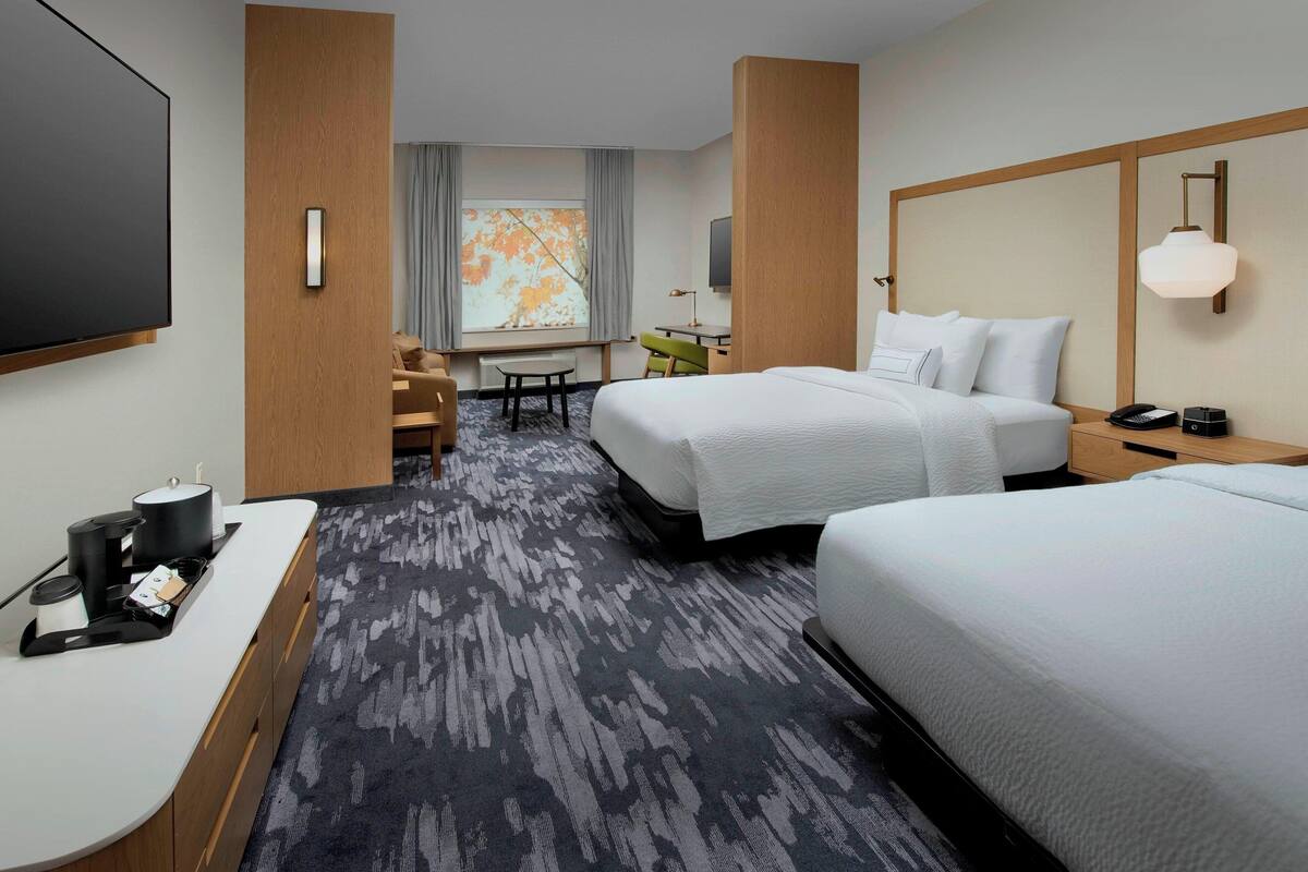Ein Hotelzimmer mit zwei Betten, einem Schreibtisch mit Stuhl, einem Fernseher, einer Lampe und einem Fenster mit Vorhängen.