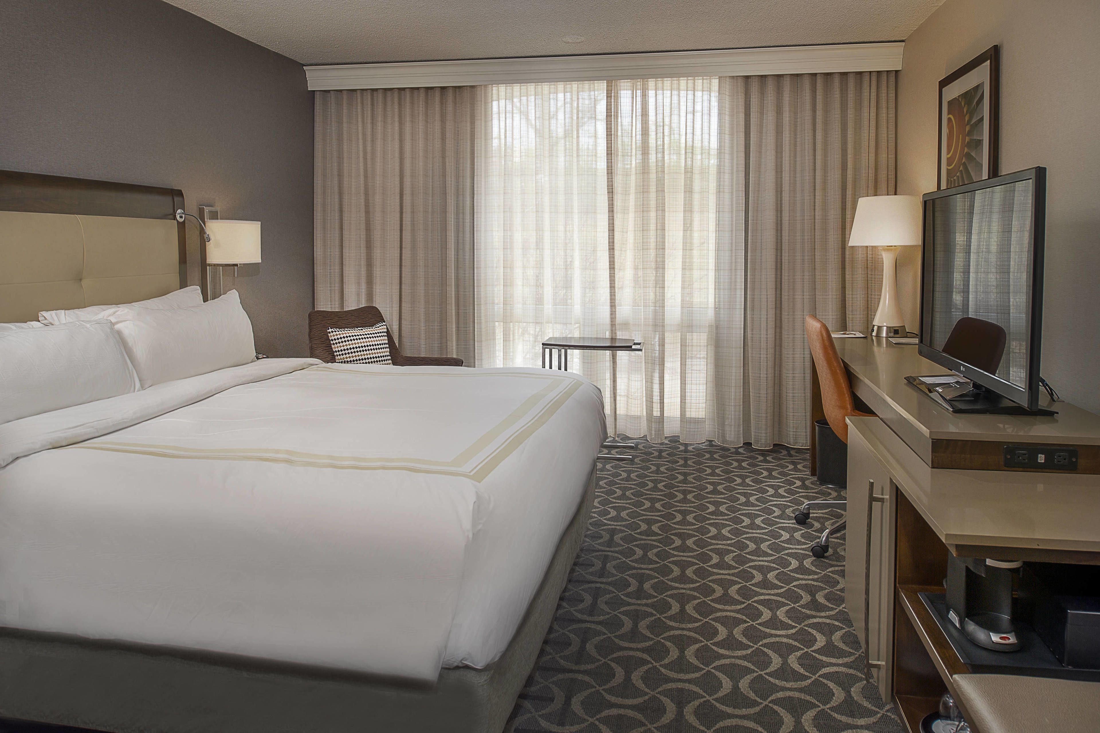 A hotel room with a large bed, a desk with a television, a chair, a small table, and a lamp.