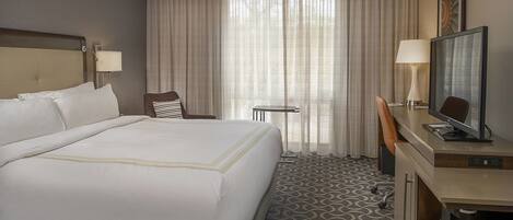 A hotel room with a large bed, a desk with a television, a chair, a small table, and a lamp.
