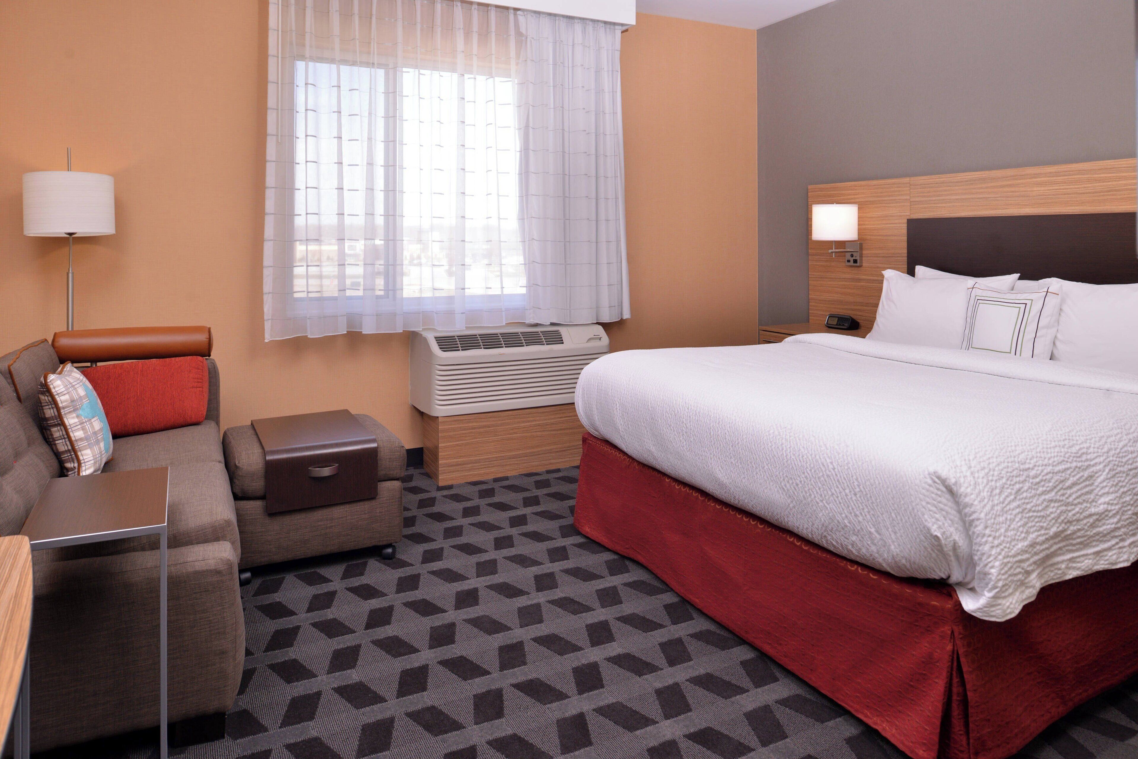Quarto de hotel com uma cama, um sofá, uma mesa pequena, uma luminária e uma janela com cortinas.