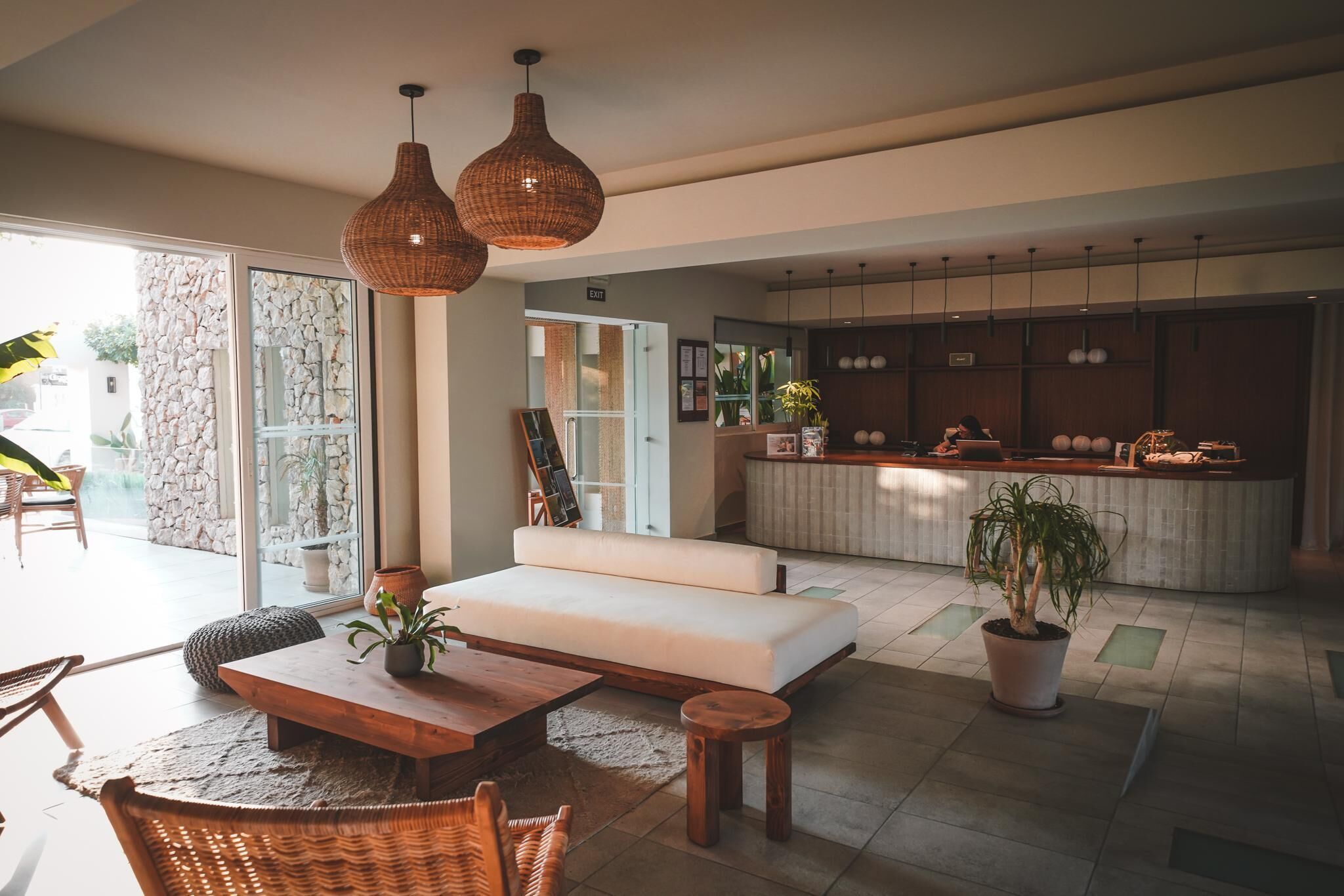 A modern living room with a sofa, wooden coffee table, and wicker chairs. There is a kitchen area with a counter and cabinets, and a large window providing natural light.