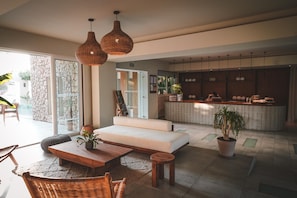 A modern living room with a sofa, wooden coffee table, and wicker chairs. There is a kitchen area with a counter and cabinets, and a large window providing natural light. - Ananea More Meni Kos ex More Meni Beach (Kos)