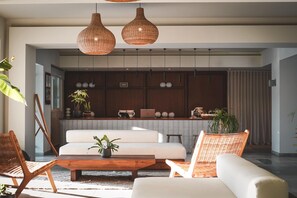 A modern living room with a sofa, two wicker chairs, a wooden coffee table, and a kitchen area with a laptop, plants, and decorative items. - Ananea More Meni Kos ex More Meni Beach (Kos)