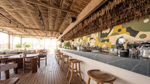 Un restaurant amb taules i tamborets de fusta, una zona de bar amb barra i un teulat de palla.