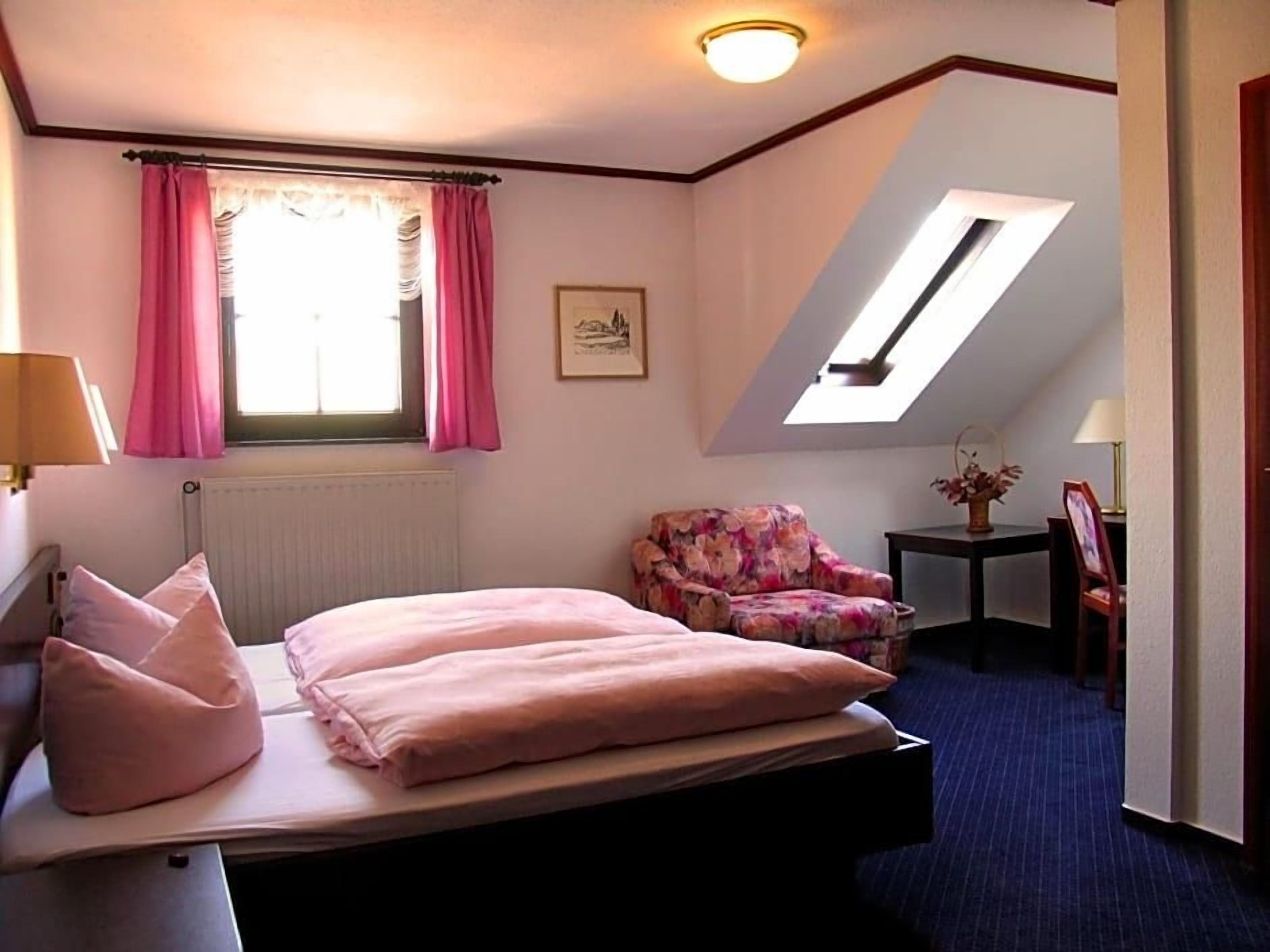 Un dormitorio con cama, una silla, un escritorio, un ventanal con cortinas y un tragaluz.