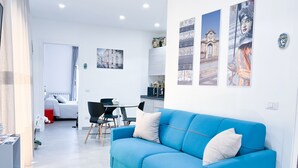 Uma sala de estar moderna com um sofá azul, um espaço de refeições com mesa e cadeiras, e uma cozinha ao fundo.