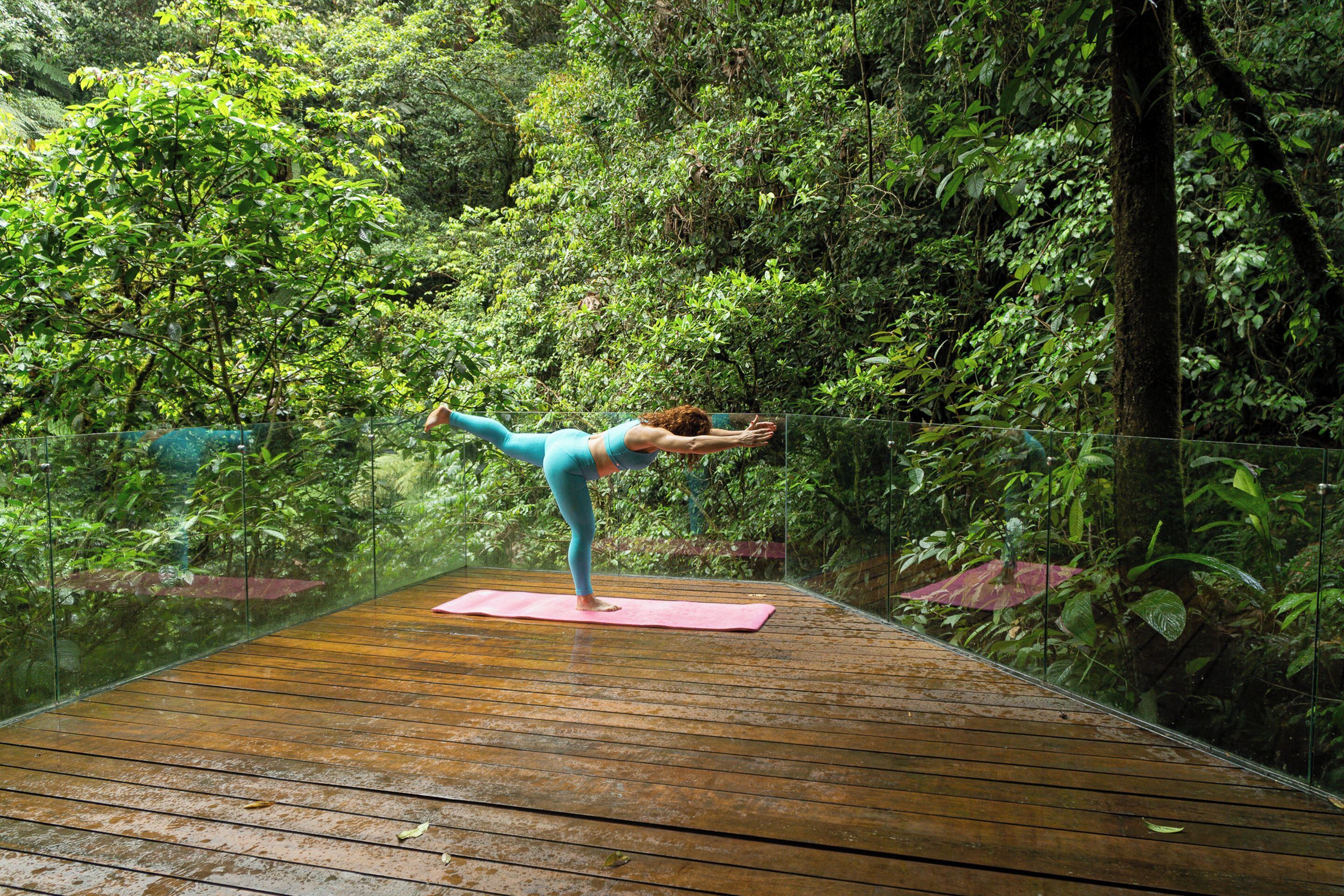 Una persona realiza una postura de yoga sobre una terraza de madera rodeada de abundante vegetación.