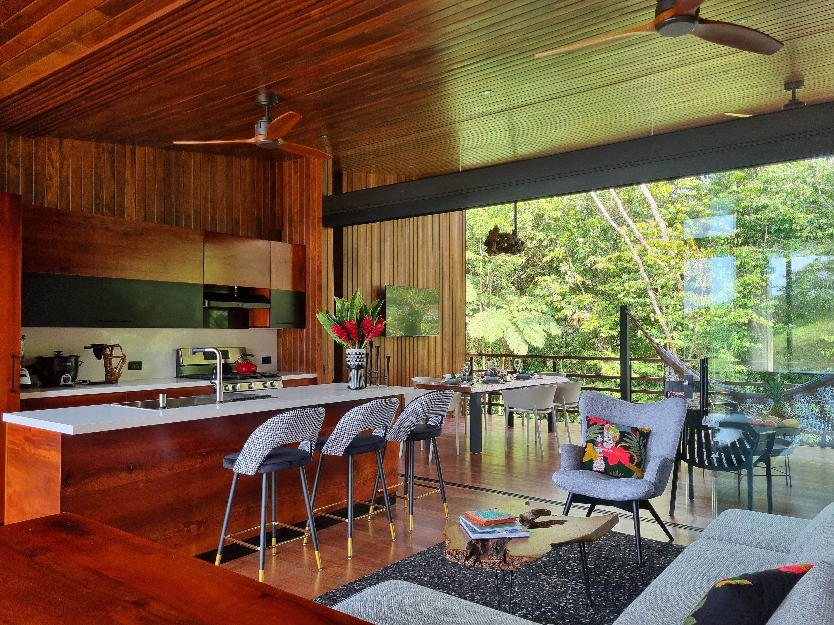 A modern kitchen with wooden cabinetry, a central island with bar stools, and a dining area with a view of lush greenery.