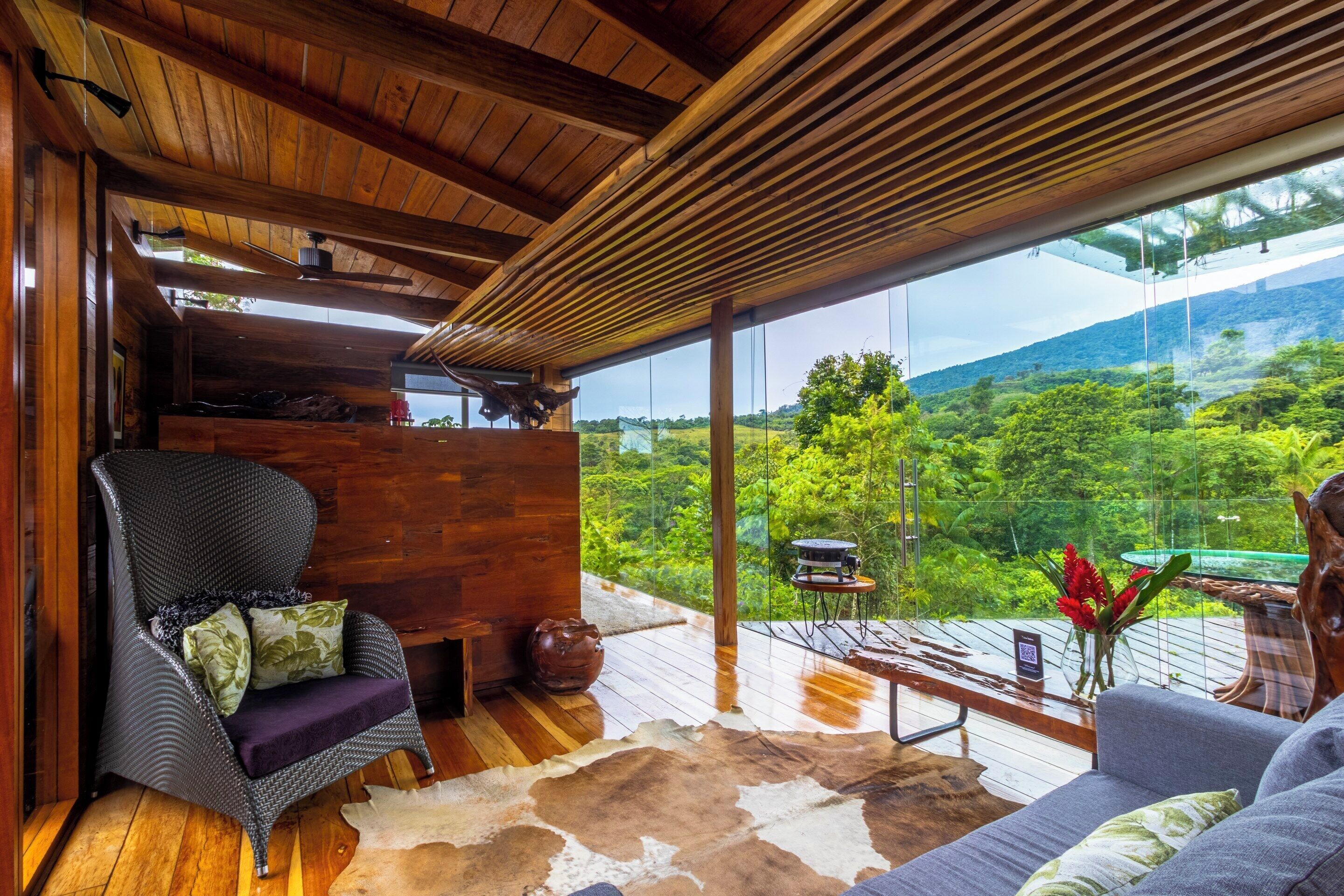 Um interior acolhedor com teto de madeira, uma janela ampla que oferece vista para vegetação exuberante, um sofá confortável e uma poltrona de vime.