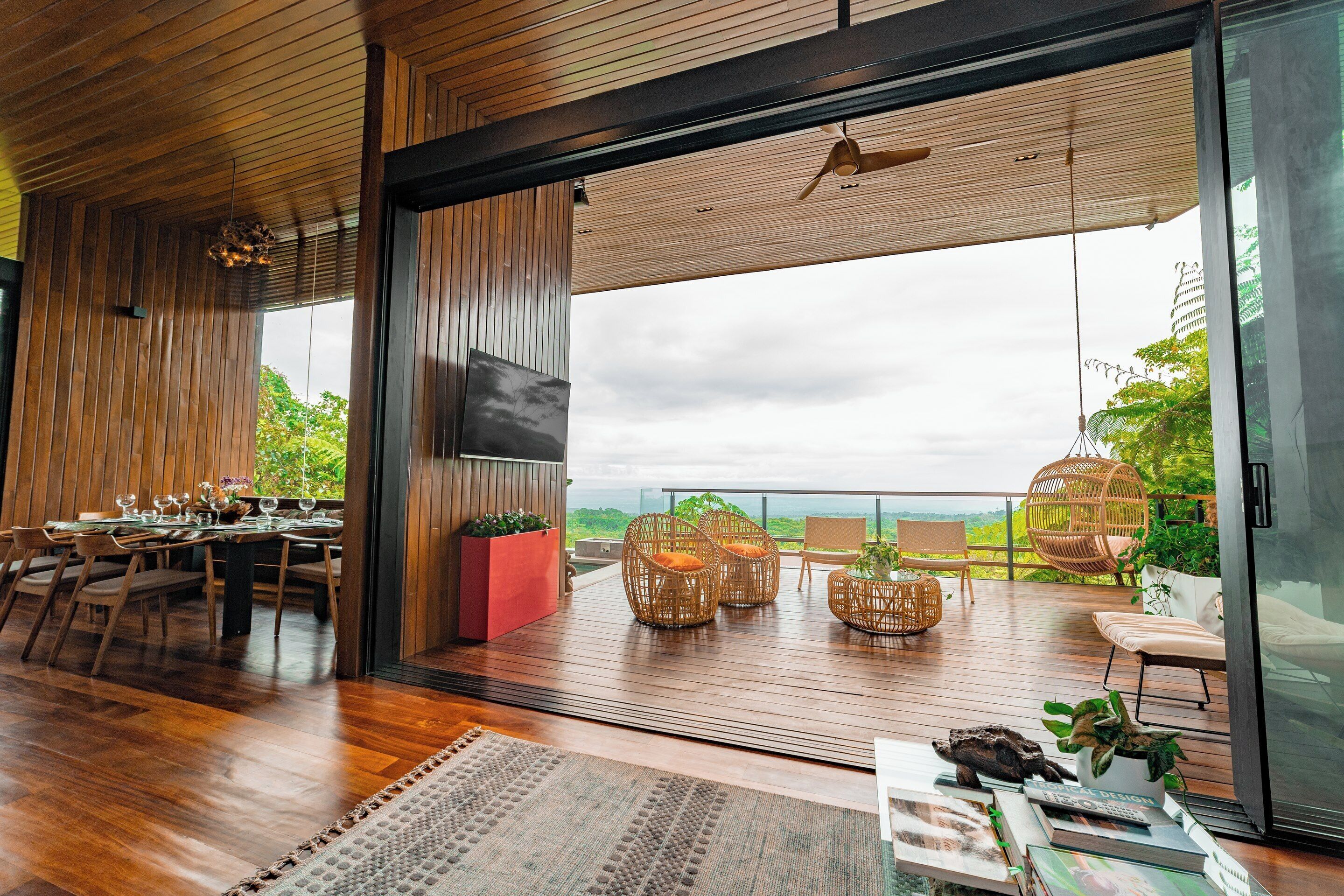 A spacious room with a wooden floor, a dining area with a table and chairs, a large window with a view of greenery, and a balcony with wicker furniture.