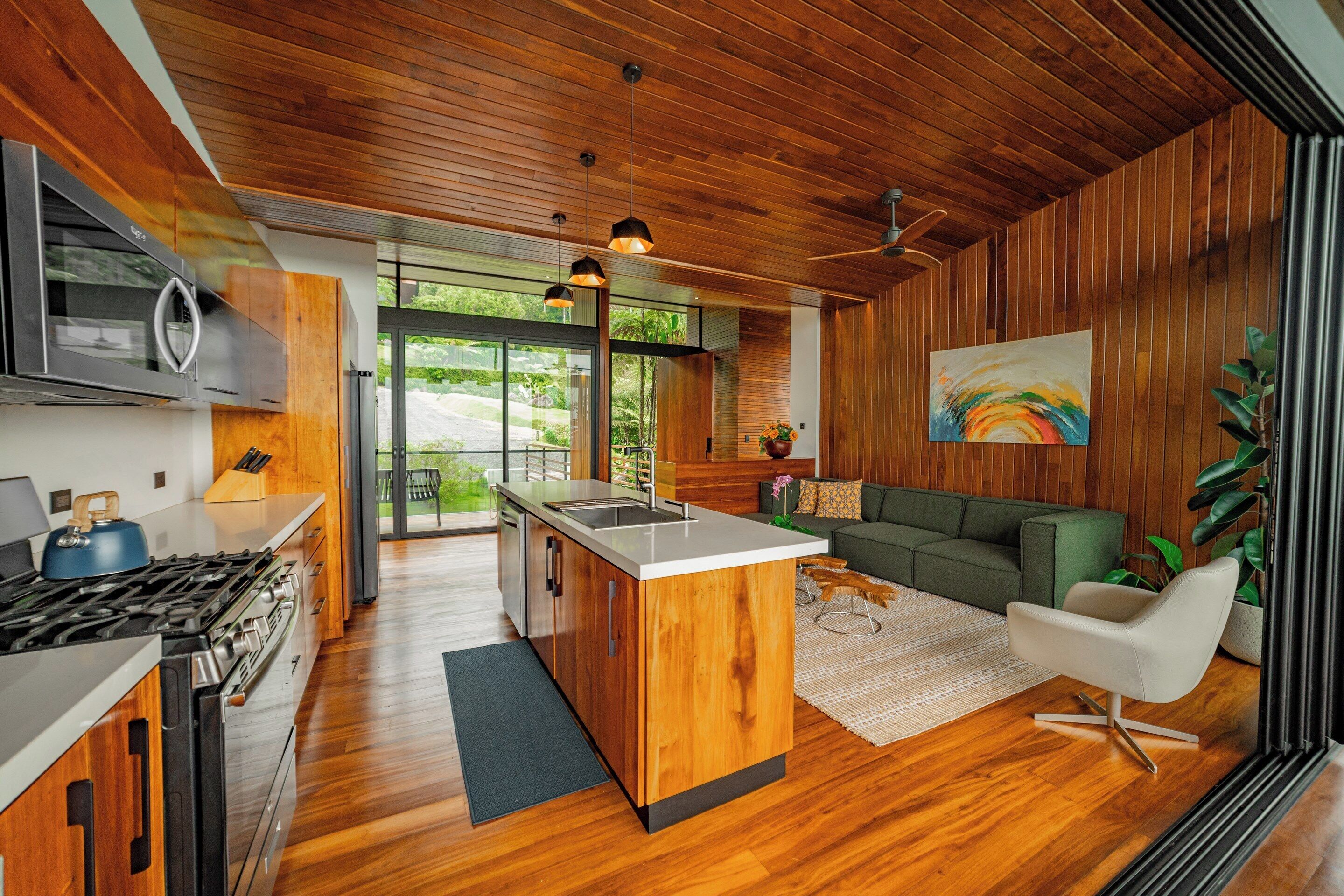 A modern kitchen with wooden cabinetry, a central island, and a dining area with a green sofa and white chair.
