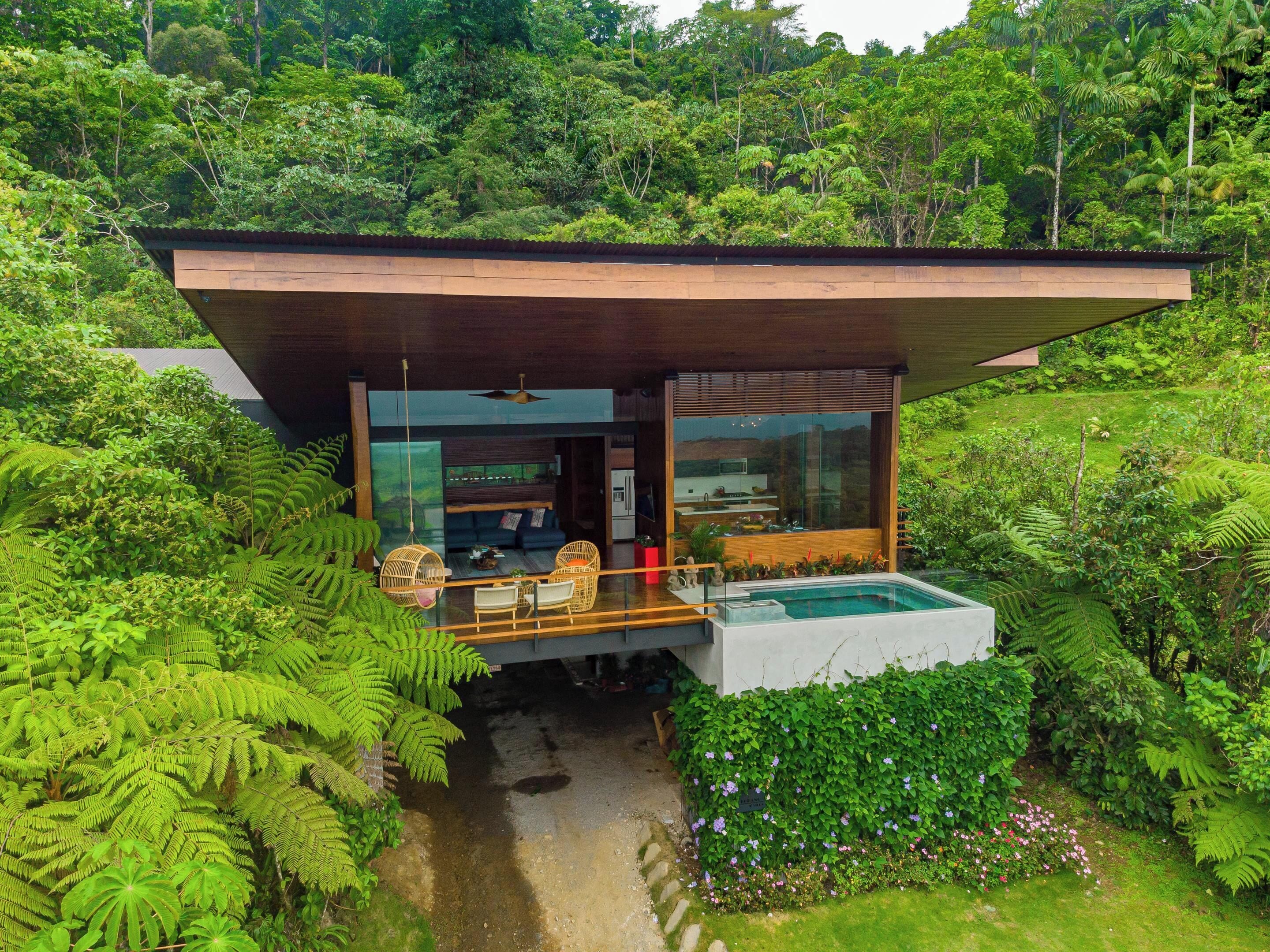 A modern house with a large glass window, a wooden deck, and a swimming pool surrounded by lush greenery.
