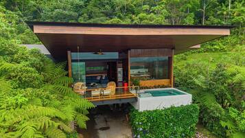 A modern house with a large glass window, a wooden deck, and a swimming pool surrounded by lush greenery.
