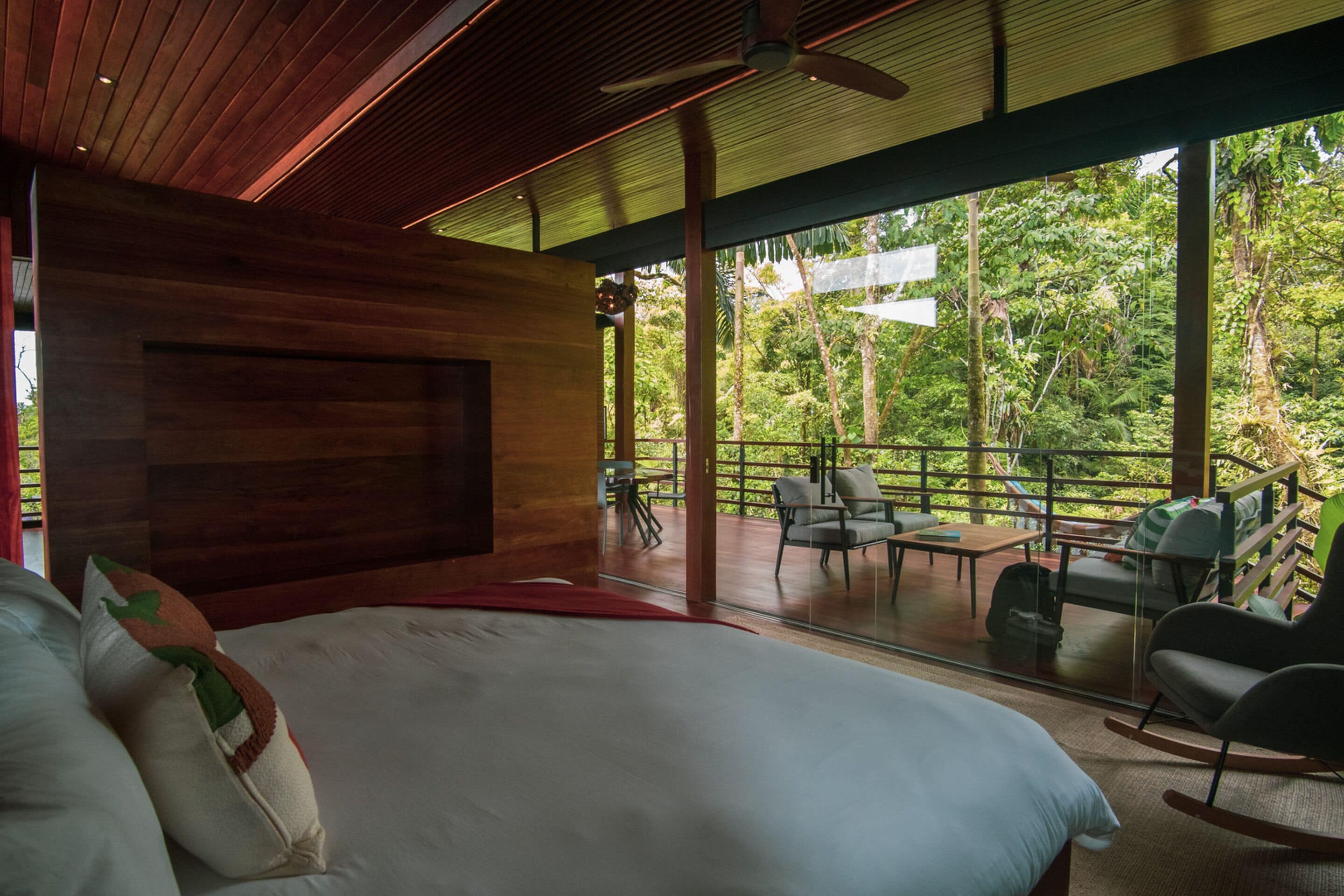 A bedroom with a large bed, wooden ceiling, and a view of a lush garden through large windows.