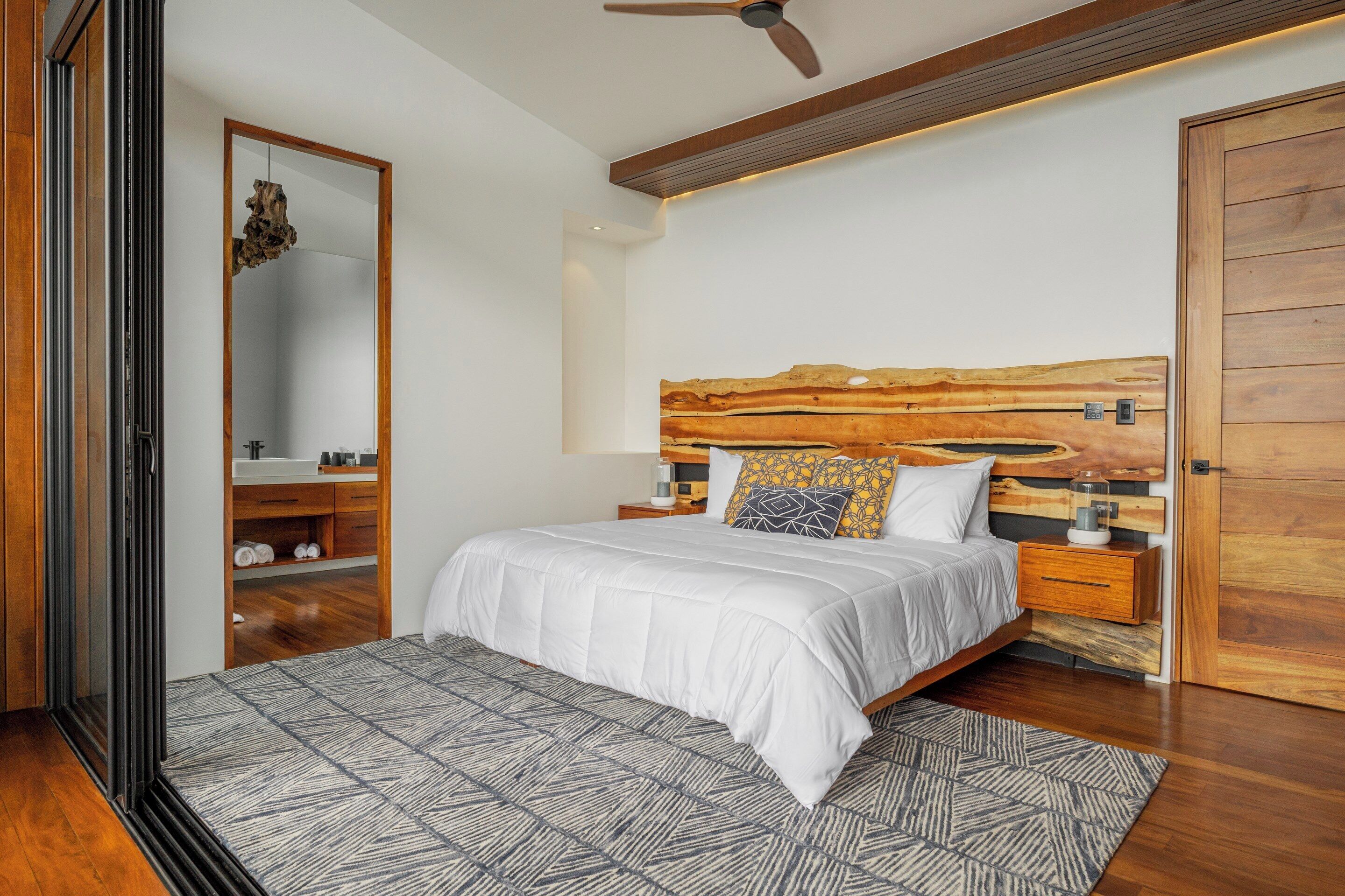 A modern bedroom with a wooden headboard, a large mirror, a bed with white bedding, and a patterned rug.