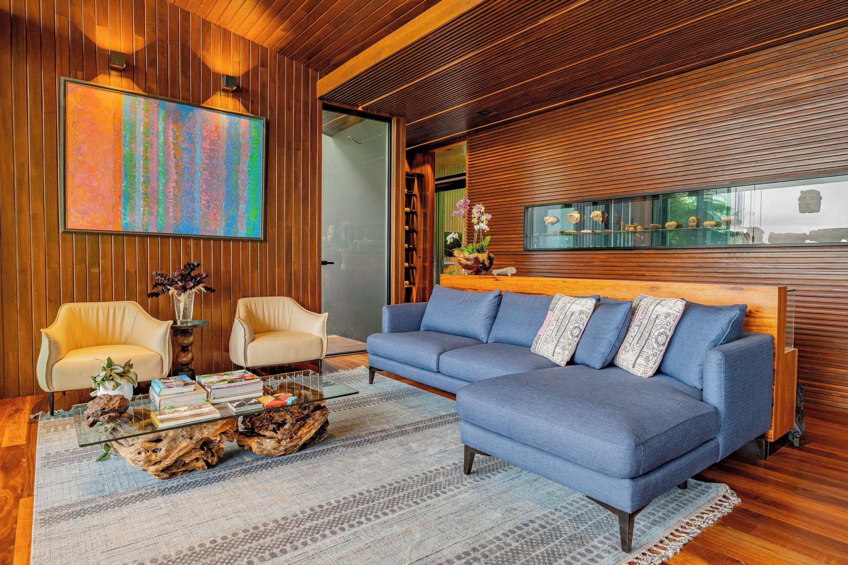 A modern living room with a blue sofa, two armchairs, a coffee table with books and a plant, and a large abstract painting.