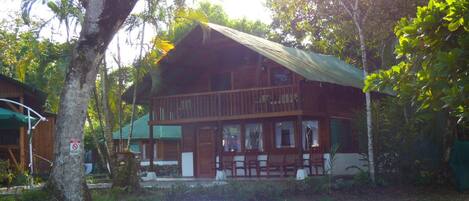 A two-story wooden cabin with a balcony, surrounded by lush greenery and a clear path leading to the entrance.