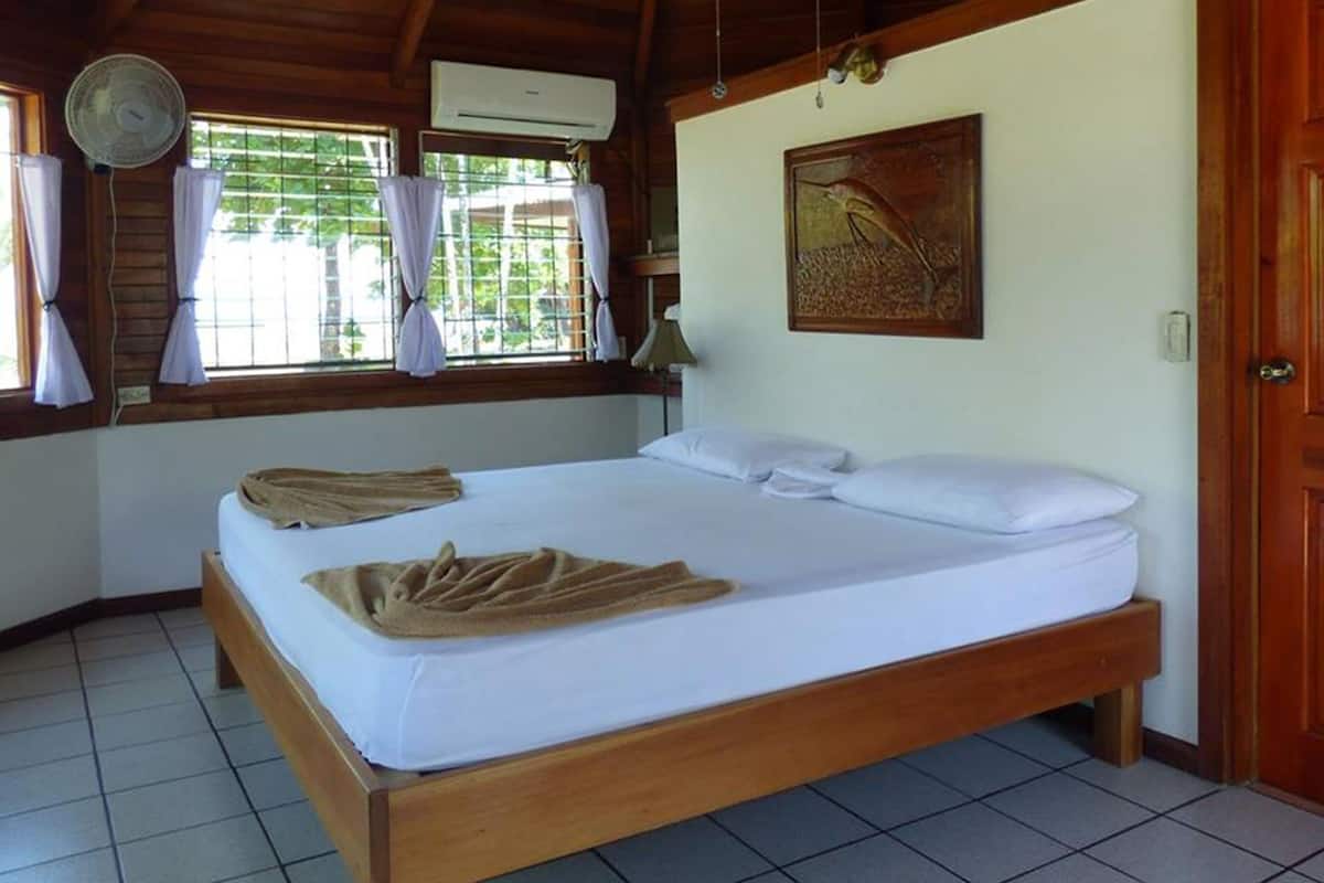 A single-bed room with a wooden ceiling, tiled floor, and a window with curtains.