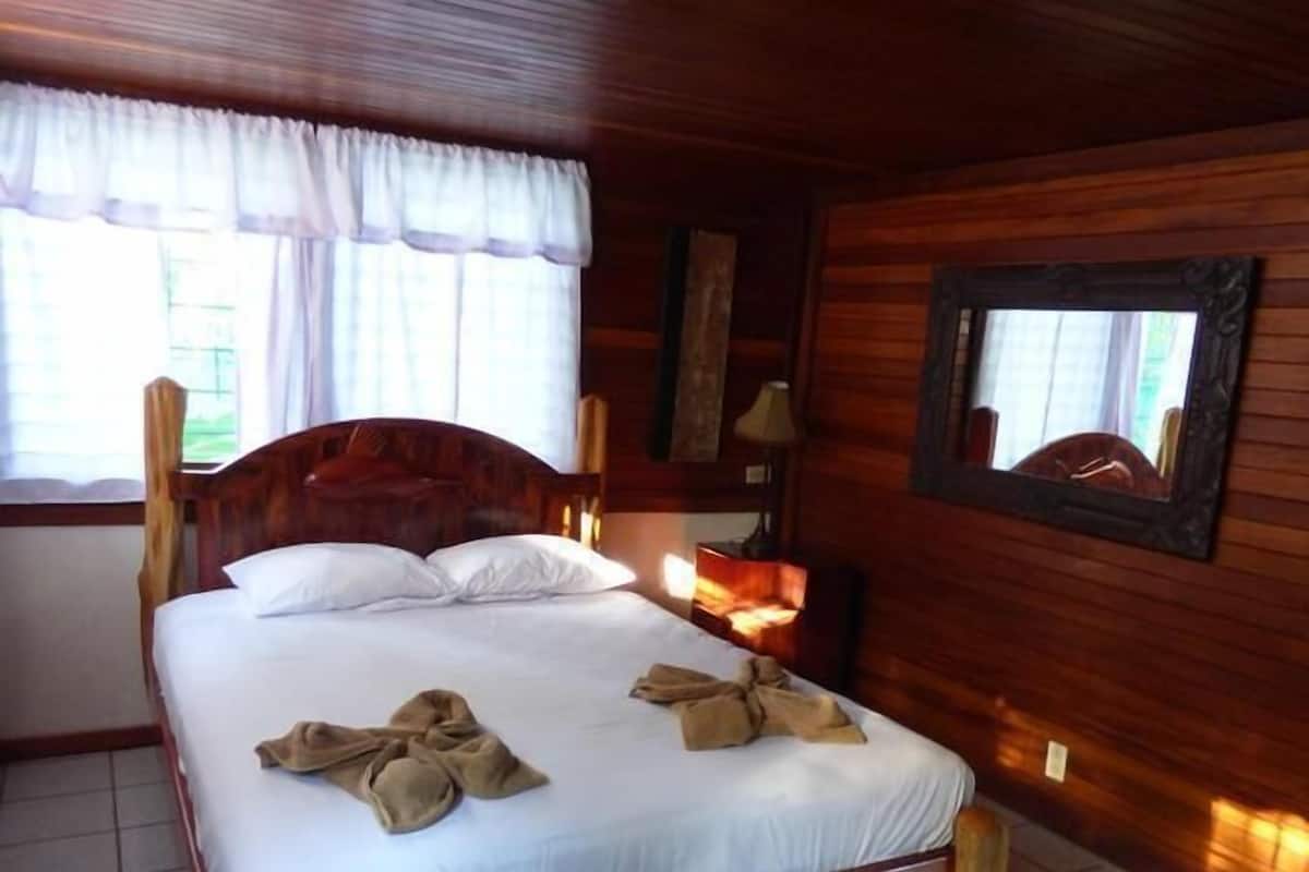 A wooden-framed bed with white linens, a bedside table with a lamp, and a window with curtains.