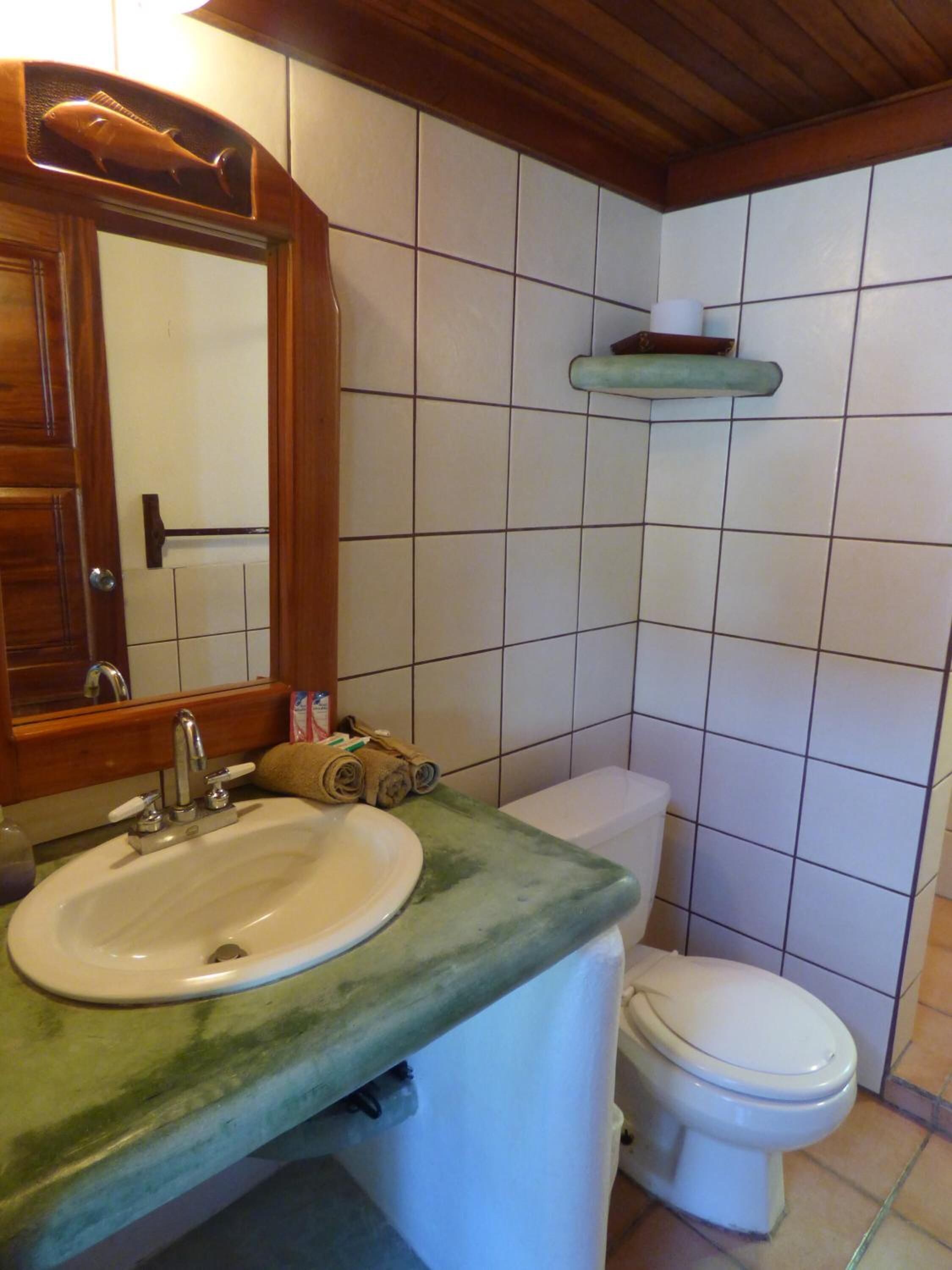 Une salle de bain avec un meuble-lavabo en bois, un lavabo blanc, un miroir, des toilettes et une étagère au-dessus des toilettes.