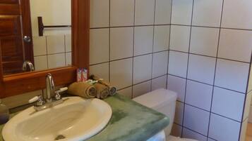 A bathroom with a wooden vanity, a white sink, a mirror, a toilet, and a shelf above the toilet.