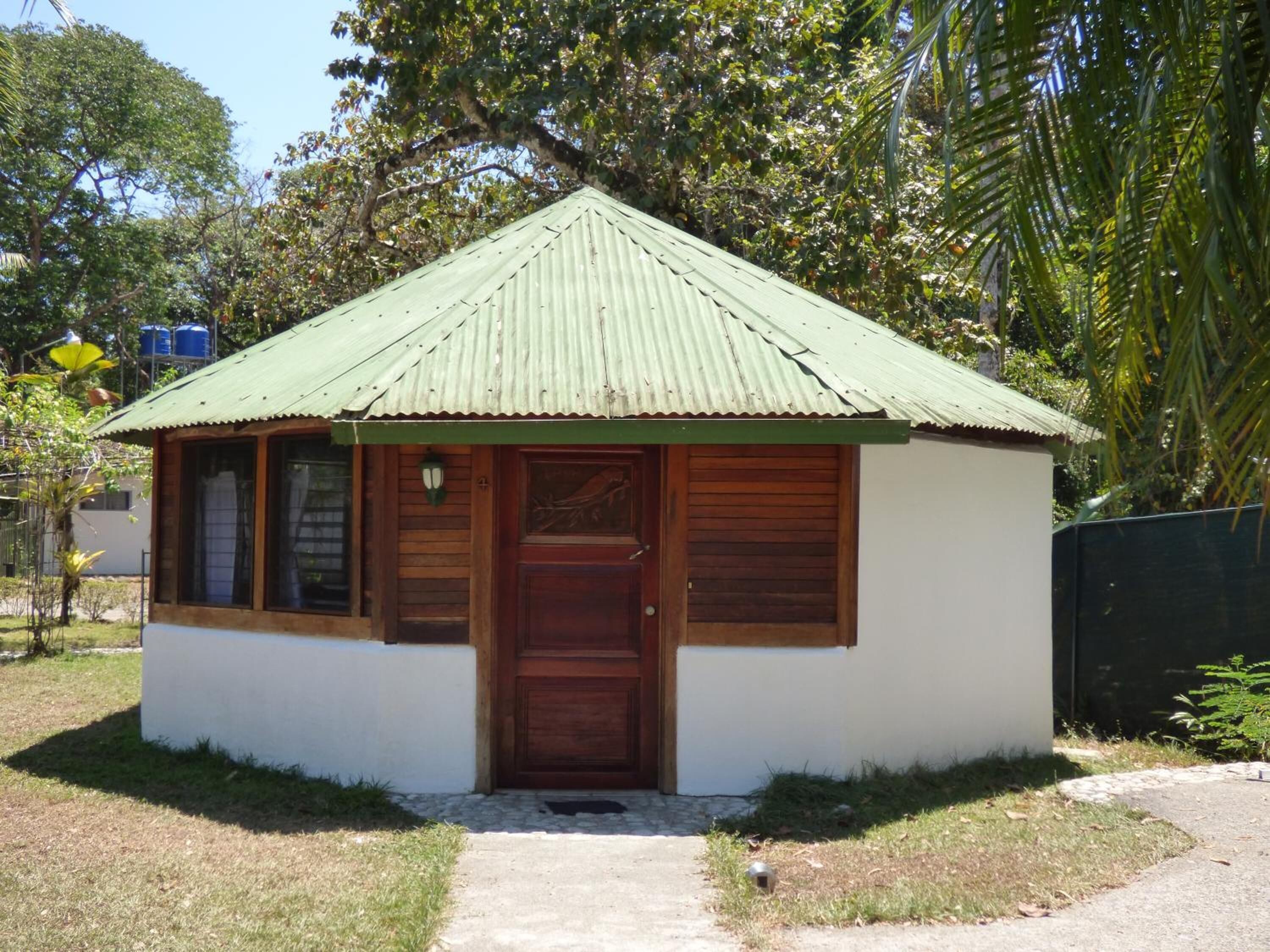 Un edificio pequeño de una sola planta, con techo de lámina verde corrugada, puerta de madera y ventana con persianas de madera.