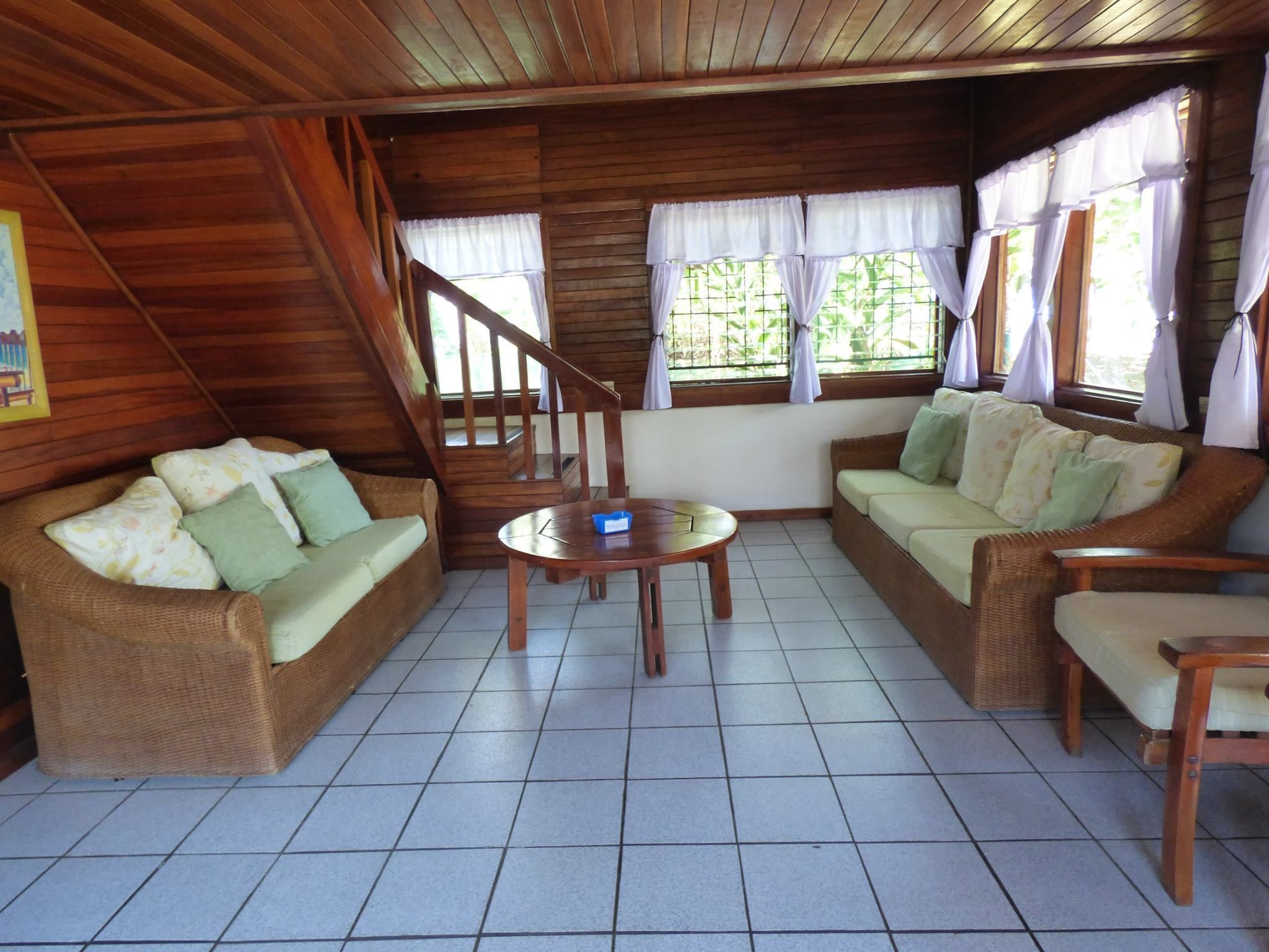 A cozy room with a wooden ceiling, a staircase, two wicker sofas, a wooden coffee table, and a window with curtains.