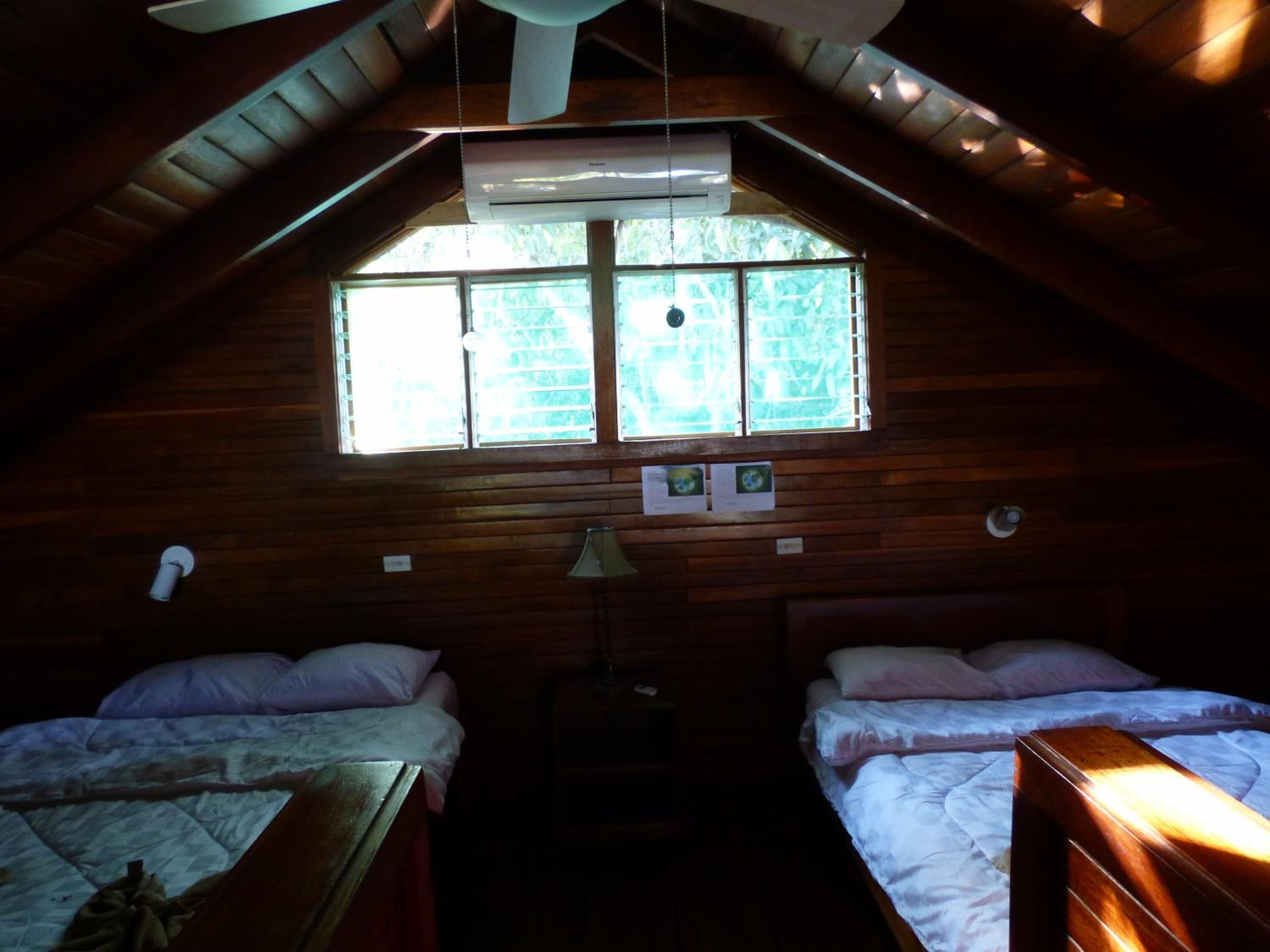 A cozy attic room with two beds, a ceiling fan, and a large window with blinds.