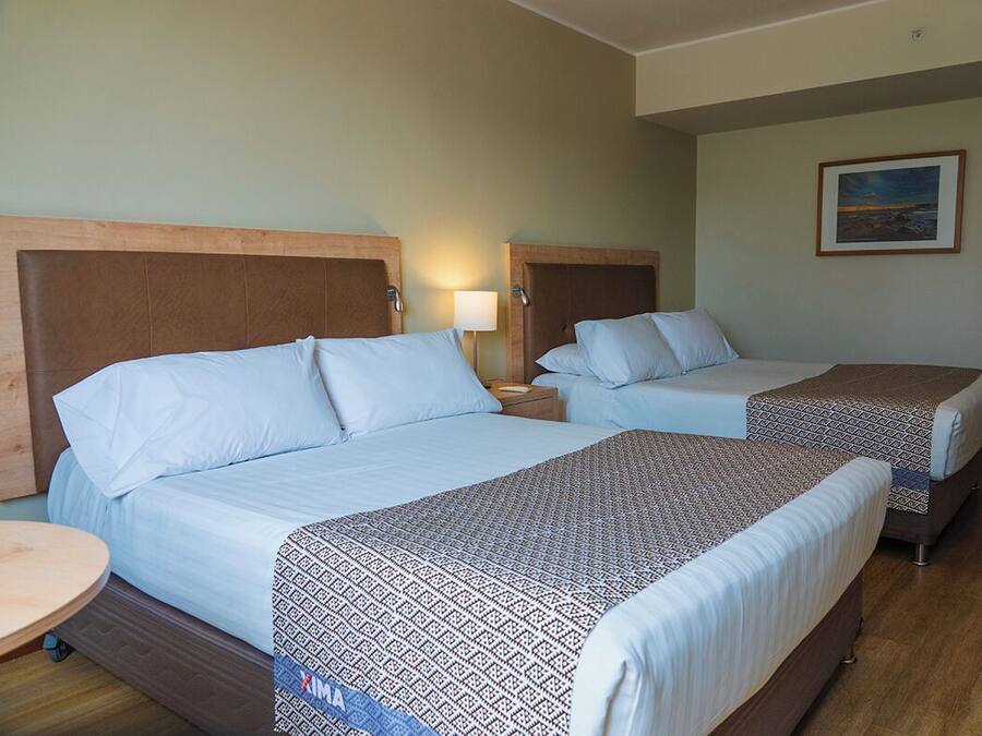 Quarto de hotel com duas camas, cada uma com cabeceira, mesa de cabeceira com abajur e um quadro na parede.