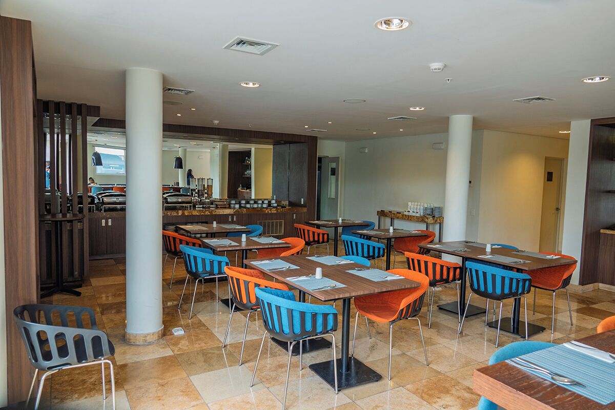 A modern restaurant with multiple tables and chairs, a bar area, and a visible kitchen in the background.