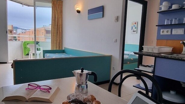 Une chambre d’hôtel avec une table à manger mise, sur laquelle on trouve un livre, une tasse, une assiette de pâtisseries et une cafetière. Une fenêtre offre une vue sur les bâtiments à l’extérieur.