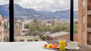 Una mesa de desayuno con fruta y jugo, vista a un paisaje urbano y montañas a través de una ventana.