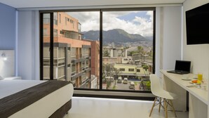 Una habitación de hotel moderna con un gran ventanal que ofrece vistas a la ciudad, una cama, un escritorio con una computadora portátil y una silla.