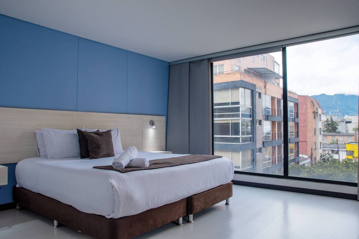 A modern hotel room with a large bed, a nightstand, a wall-mounted lamp, and a large window offering a city view.
