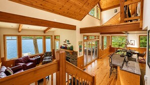 Un interior espacioso con una escalera de madera, un comedor y grandes ventanales con vistas al agua.