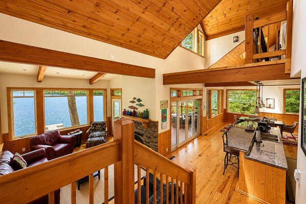 A spacious interior with a wooden staircase, a dining area, and large windows overlooking a body of water.