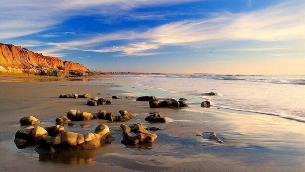 Una playa con rocas grandes en la arena y acantilados al fondo.