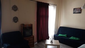 A living room with a dark blue sofa, a wooden TV stand with a television, a small table, and a window with red curtains. - The bohemian studio by the center (Thessaloniki)