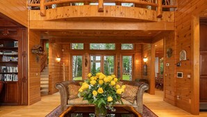 A spacious log cabin interior with a central seating area, a wooden staircase, and large windows offering views of the surrounding forest.